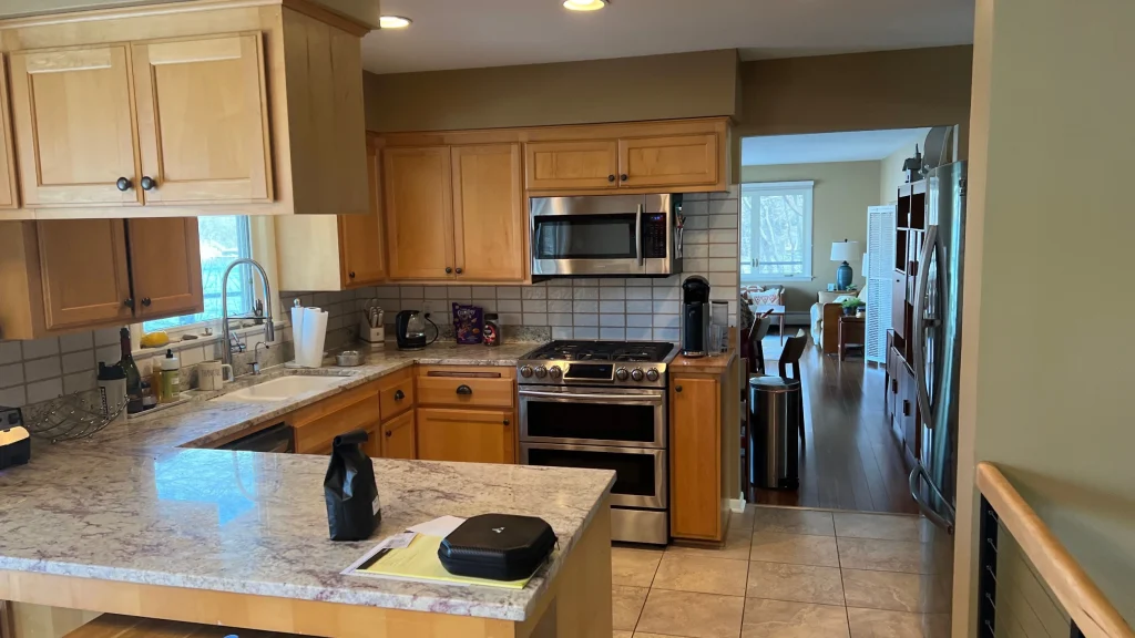 Modern kitchen with wooden cabinets, stainless steel appliances, granite countertops, and tile floor, opening into a living area with wood flooring and large windows.