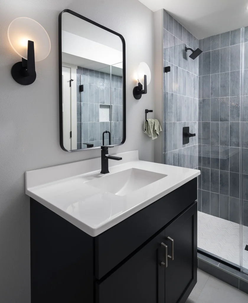 Modern bathroom with a black vanity, rectangular mirror, wall sconces, and a walk-in shower with gray tiles and black fixtures.