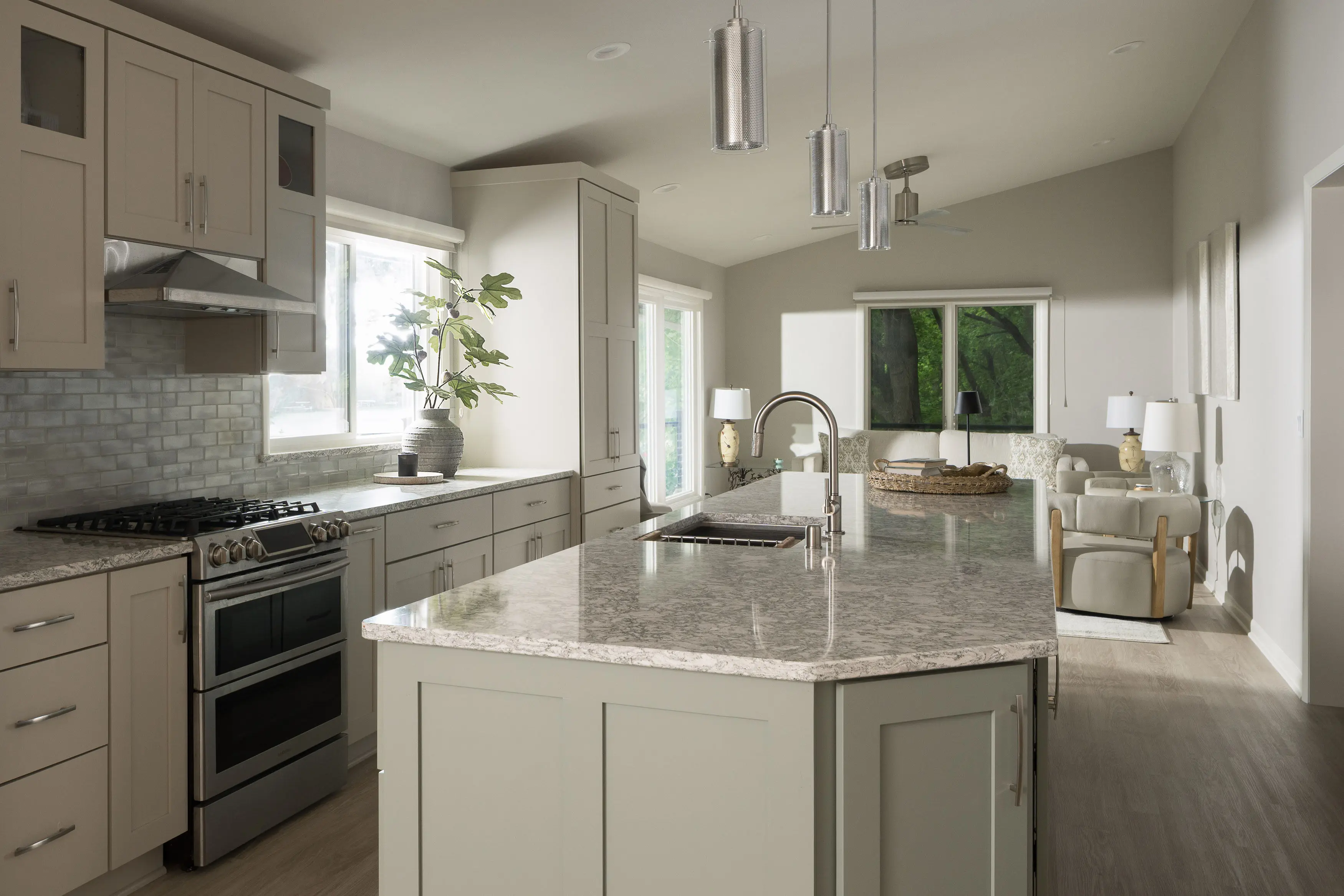 Modern kitchen with light gray cabinets, stainless steel appliances, marble-patterned island, pendant lights, and an open view into a living area with large windows.